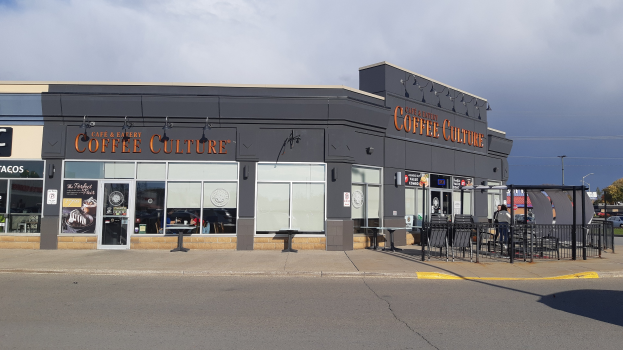 Ein Kaffee-Shop an einer Straßenecke mit einer Straße im Vordergrund, einem Gebäude im Hintergrund, Fahrzeugen, Bäumen und einem blauen und weißen Himmel.