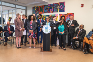 Gruppe von Menschen vor einem Rednerpult mit Mikrofon und einer Tafel mit dem Text 'Sancho City Councilman Joins City Council' stehend, mit sitzenden und stehenden Zuschauern, Flaggen, Feuerlöscher und Deckenleuchten im Hintergrund.