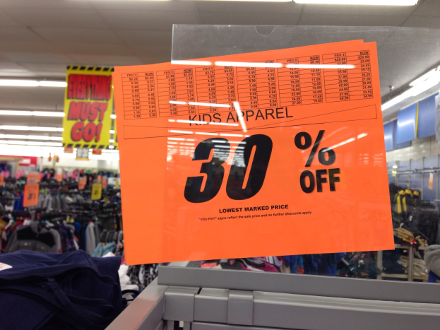 Ladenfront mit einem "Kids Apparel 30% Off"-Schild im Vordergrund, Kleidung an Bügeln im Hintergrund, Deckenleuchten und eine Tafel mit Text auf der rechten Seite.