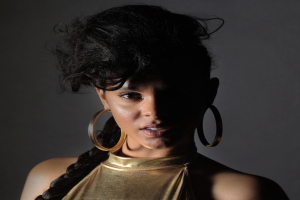 Eine Frau in einem goldenen Kleid und großen Kreolen steht selbstbewusst vor einer Wand, ihr glatter Bob und natürliches Make-up unterstreichen ihren entschlossenen Ausdruck, während sie direkt in die Kamera schaut.