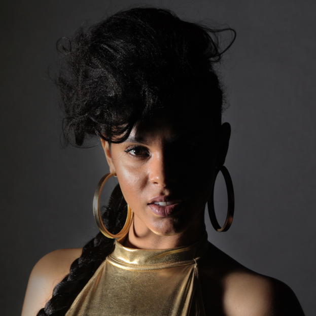 Eine Frau in einem goldenen Kleid und großen Kreolen steht selbstbewusst vor einer Wand, ihr glatter Bob und natürliches Make-up unterstreichen ihren entschlossenen Ausdruck, während sie direkt in die Kamera schaut.