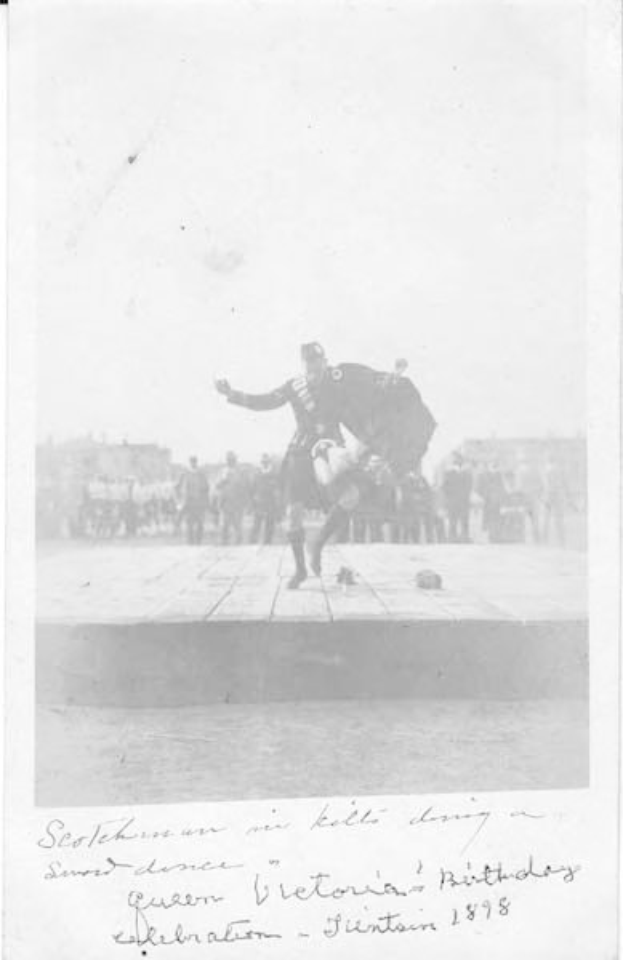 Schwarz-weißes Foto eines Mannes in einem Kilt, der auf einer Bühne tanzt, während der Geburtstagsfeier von Königin Victoria in Edinburgh 1898, mit einer Menge und Gebäuden im Hintergrund.