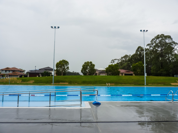Große rechteckige Schwimmbad in einem Park, umgeben von Zäunen, Pfosten und Lichtern mit Bäumen drumherum, Häuser im Hintergrund unter einem klaren blauen Himmel.