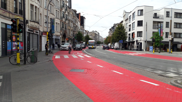 Rote Fahrradspur neben einer Stadtstraße mit fahrenden Autos, Fußgängern auf dem Gehweg, Gebäuden mit Fenstern, Bäumen und einem klaren blauen Himmel im Hintergrund.