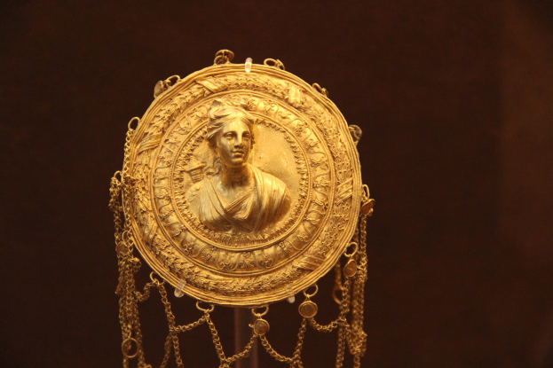 Gold medallion mit einer Frauengestalt, das mit detaillierten Verzierungen und einem luxuriösen Design versehen ist und vor einem dunklen Hintergrund zu sehen ist.