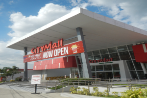 Ein belebtes Stadtzentrum mit einer Tafel "City Mall Now Open" umgeben von Gebäuden, Schildern, Geländern, Pflanzen, Bäumen, Strommasten mit Drähten und einem bewölkten Himmel im Hintergrund.