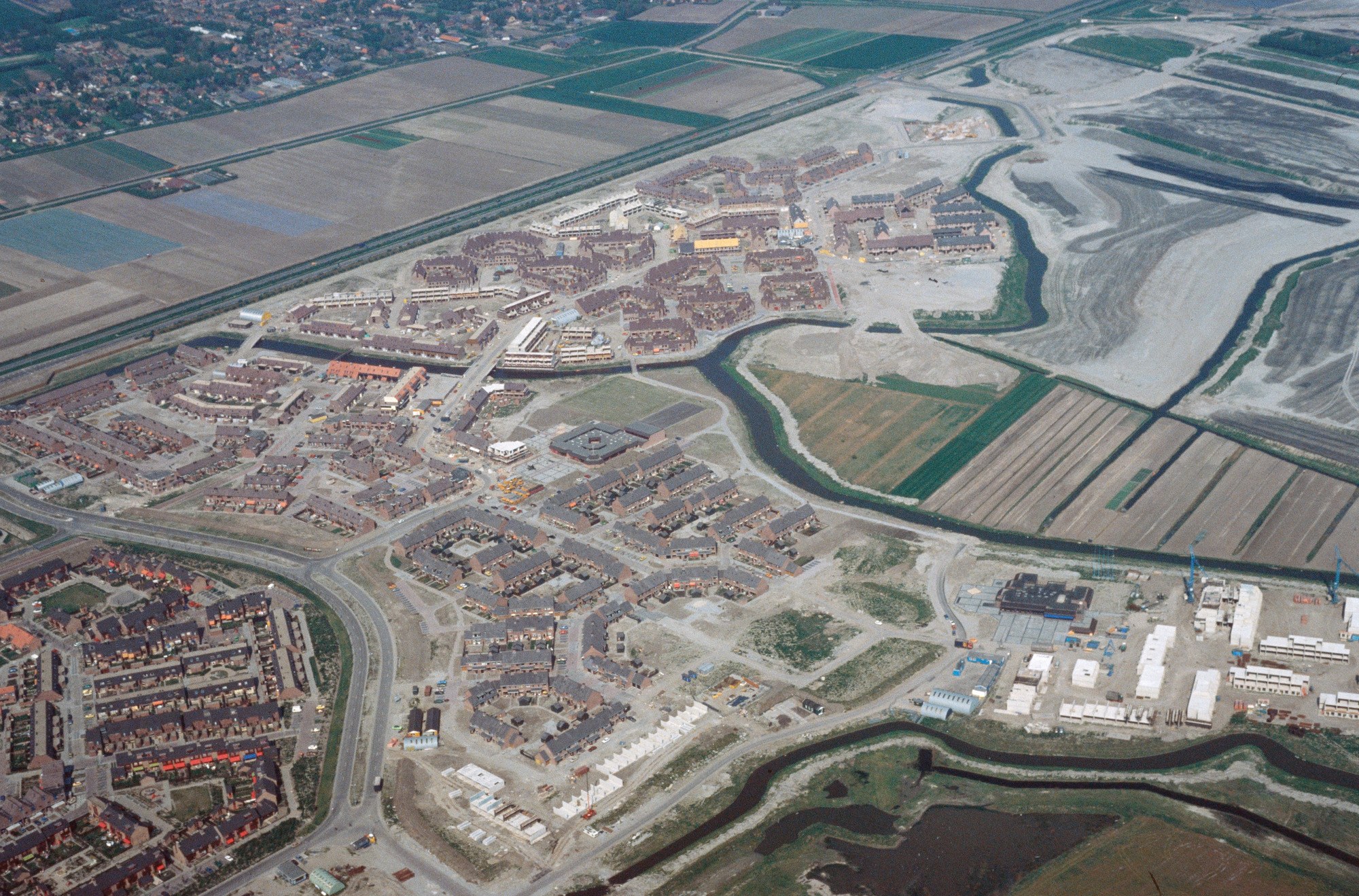 Luftaufnahme einer Stadt mit dichtem Baumbestand, Straßen, Feldern, Bäumen und einem zentralen Reservoir, umgeben von städtischen und grünen Bereichen.
