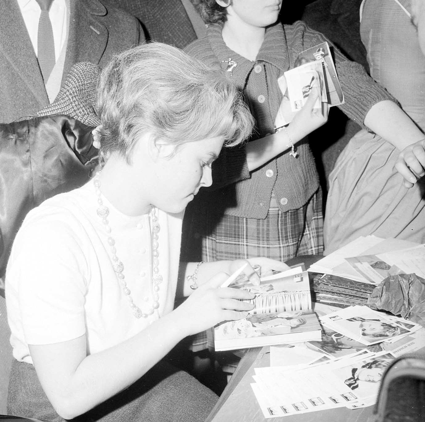 Schwarz-weiß-Foto von Prinzessin Diana, die an einem Tisch sitzend ein Buch liest, umgeben von einer Gruppe von Menschen.