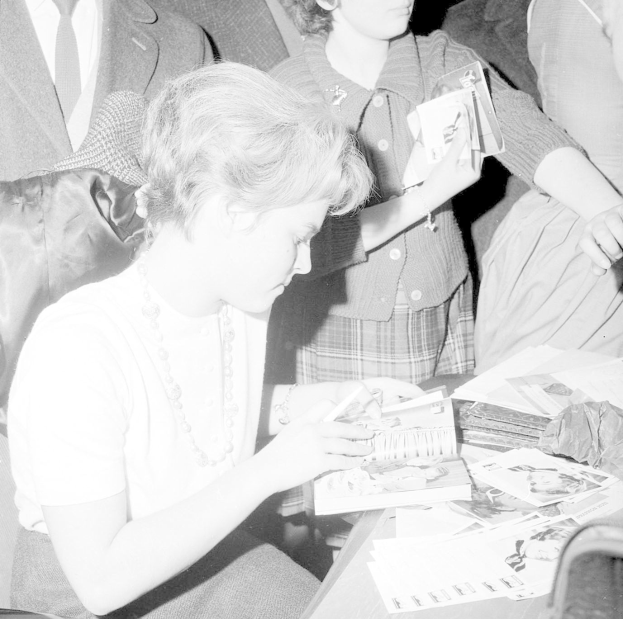 Schwarz-weiß-Foto von Prinzessin Diana, die an einem Tisch sitzend ein Buch liest, umgeben von einer Gruppe von Menschen.