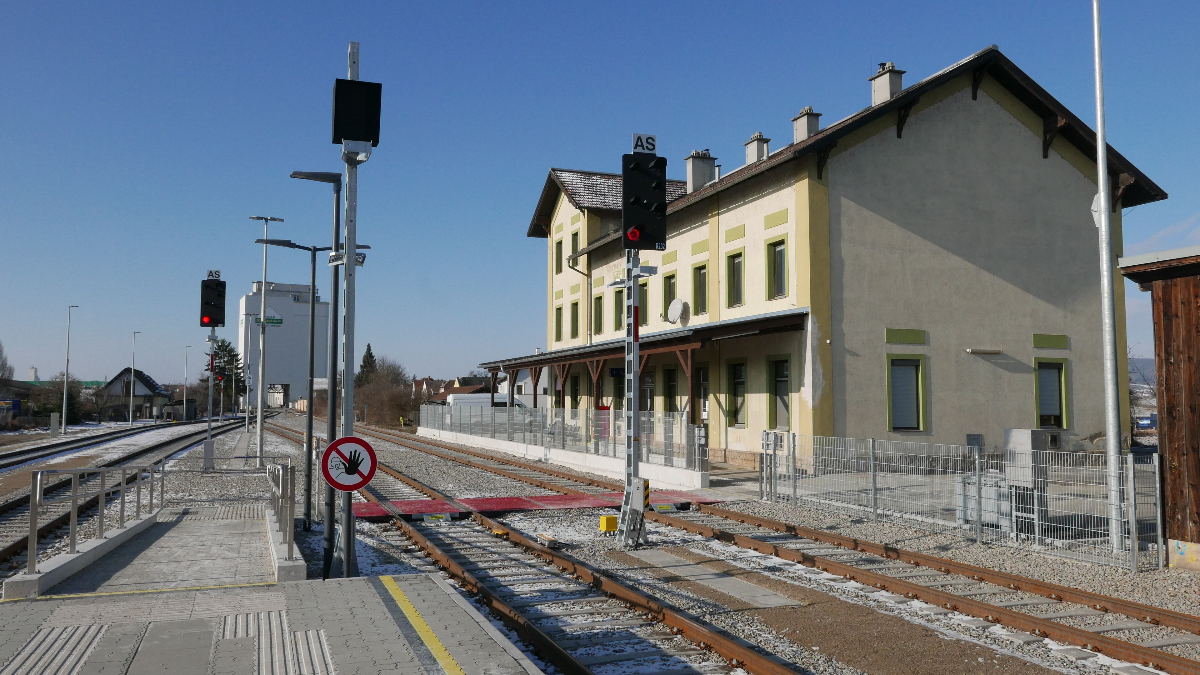 Ein belebter Bahnhof mit einem Zug auf den Gleisen, umgeben von Gebäuden, Straßeninfrastruktur und Bäumen unter einem klaren blauen Himmel.