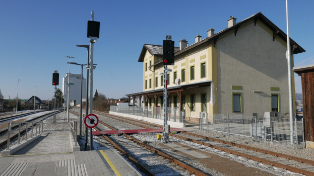 Ein belebter Bahnhof mit einem Zug auf den Gleisen, umgeben von Gebäuden, Straßeninfrastruktur und Bäumen unter einem klaren blauen Himmel.