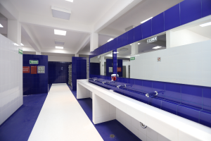 Ein öffentliches WC mit blauen Fliesenwänden und weißen Arbeitsplätzen, mit Waschbecken mit Hähnen, einer Plattform mit einer Flasche und anderen Gegenständen, Texttafeln an den Wänden, Deckenleuchten und Säulen.