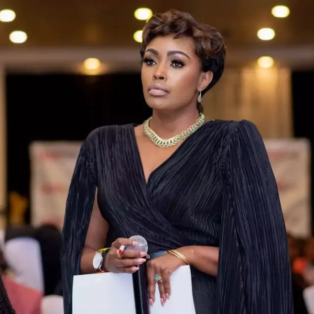 Eine Frau in einem schwarzen Kleid hält ein Mikrofon und ein Blatt Papier und spricht zu einer sitzenden Gruppe unter Deckenbeleuchtung, wobei sie in eine Diskussion vertieft zu sein scheint.