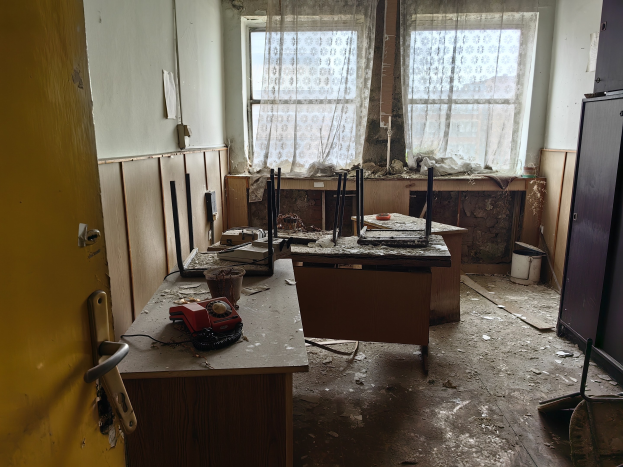 Leerer Raum mit einer Tür auf der linken Seite, einem Schrank auf der rechten Seite und einem Fenster mit Vorhängen im Hintergrund; ein Tisch in der Mitte hält verstreute Gegenstände, was auf ein verlassenes Büro hinweist.