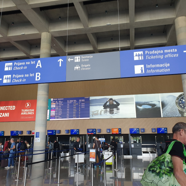 Eine Gruppe von Menschen mit Gepäck steht in einem Flughafen-Terminal in der Nähe eines "Check-in"-Schildes, mit Pfosten, Bändern, Tafeln, Säulen und Deckenleuchten im Hintergrund.