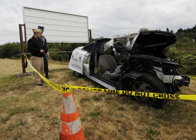 Ein Polizist in Uniform steht neben einem schwer beschädigten Auto am Straßenrand mit eingedellter Haube und zersplitterten Fenstern neben einem Verkehrskegel und einer Schnur an einem Pfahl, mit Bäumen und einem bewölkten Himmel im Hintergrund.