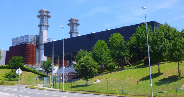 Großes Industriegebäude mit vielen Fenstern, als Kraftwerk identifiziert durch sichtbare Beschriftung, umgeben von städtischer Infrastruktur einschließlich Straßenlaternen, Fahrzeugen und Grün unter einem bewölkten Himmel.