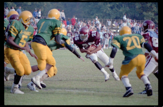 Football player in motion with the ball, surrounded by opposing team members in helmets, with spectators and trees in the background.