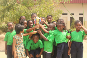 Eine Gruppe von Kindern hält einen Pokal in einem Schulhof mit Bäumen und einem Gebäude im Hintergrund.