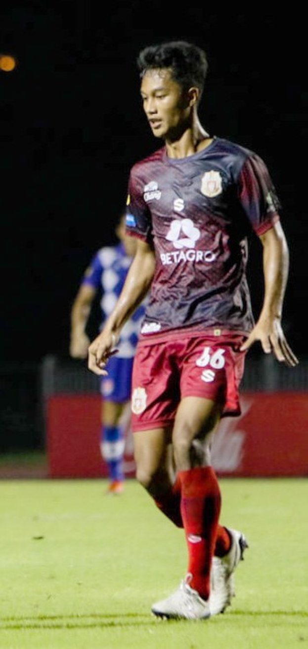 Ein Mann in einem roten Hemd und Shorts, der Fußball auf einem Feld spielt, mit einer anderen Person im Hintergrund und einer Tafel mit Text.