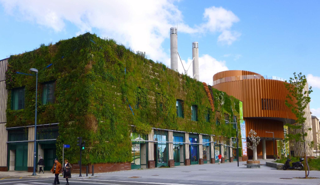 Ein großes Gebäude mit einer grünen Wand an seiner Seite, umgeben von Straßeninfrastruktur, Fahrzeugen, Fußgängern, Bäumen und einem bewölkten Himmel.