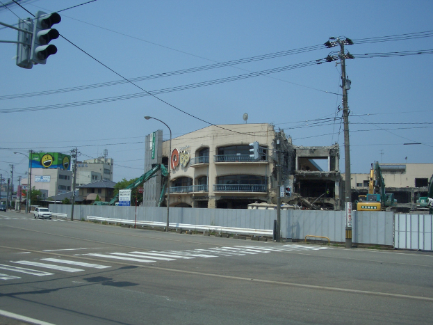 Eine Stadtstraße mit fahrenden Fahrzeugen, Strommasten, Laternen und einer Ampel auf der linken Seite, mit Gebäuden, Bäumen und einem teilweise abgerissenen Gebäude im Vordergrund.