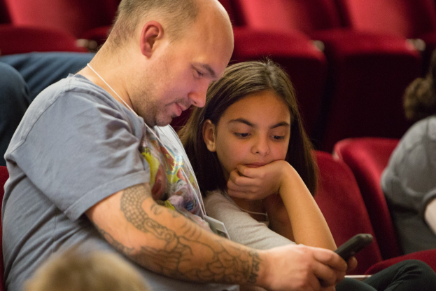 Zwei Menschen, ein Mann und ein junges Mädchen, sitzen in einem Kino, beide konzentrieren sich auf ein Handy, mit anderen Sitzenden im unscharfen Hintergrund.