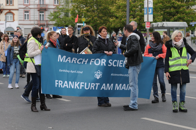 Eine bunte Gruppe von Menschen marschiert die Straße entlang, trägt ein "March for Science Frankfurt am Main"-Schild, mit Bäumen, Gebäuden und einem klaren Himmel im Hintergrund.