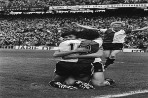 Drei Fußballspieler liegen sich auf dem Boden in den Armen nach einem Tor, mit jubelnder Menge und schwenkenden Fahnen im Hintergrund, in einer schwarz-weißen Stadionkulisse.