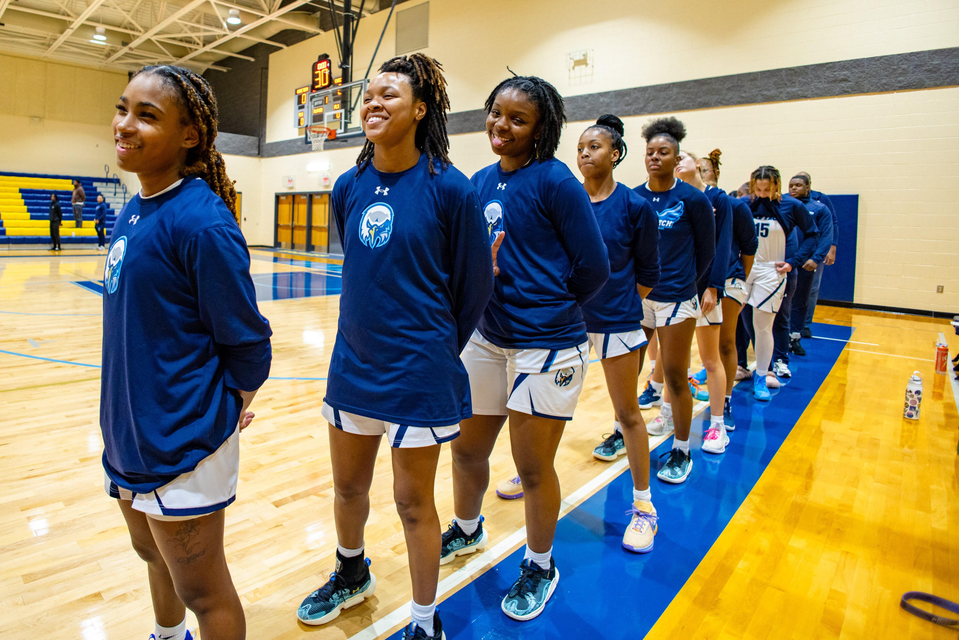 Eine Gruppe junger Frauen in blauen T-Shirts steht lachend auf einem Basketballfeld, mit Flaschen und Gegenständen rechts daneben und Zuschauern im Hintergrund.