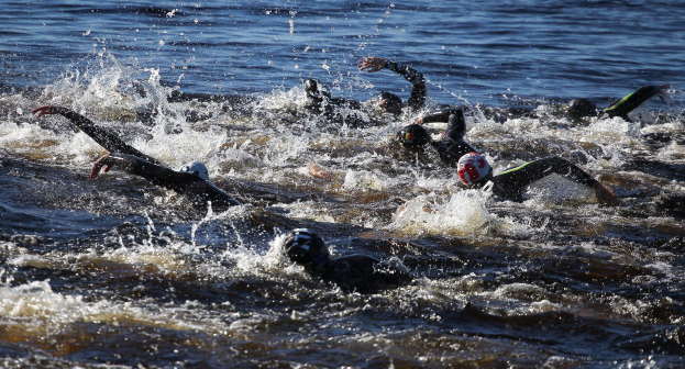 Eine Gruppe von Menschen in schwarzen Neoprenanzügen und Helmen schwimmt synchron während eines Triathlons, wobei sich die Sonne im Wasser spiegelt.