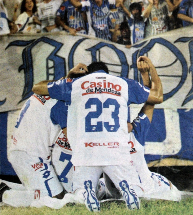 Eine Gruppe von Fussballspielern feiert ein Tor auf dem Boden vor einer jubelnden Menge, mit einem Banner mit Text im Hintergrund.