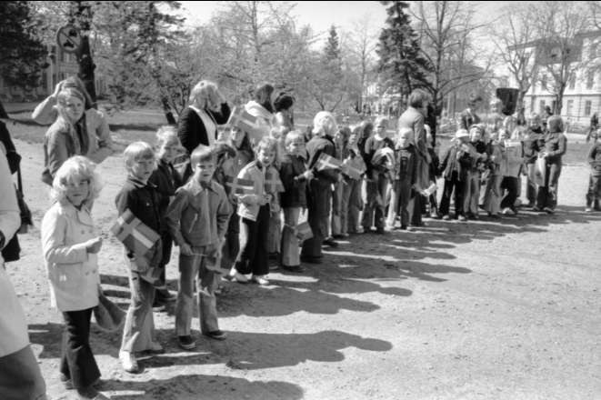 Schwarz-weißes Bild einer Gruppe von Menschen, die Fahnen auf einer Schotterstraße während einer Protestmarsch an einer Schule halten, mit Bäumen, Gebäuden und einem klaren Himmel im Hintergrund.