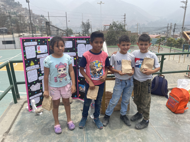 Gruppe von Kindern auf einem Basketballfeld mit Papiertüten in den Händen, Tüten und Gegenstände rechts und eine Tafel mit Text links, vor dem Hintergrund von Zäunen, Pfosten, Laternen, Strommasten, Bäumen, Gebäuden, Hügeln und Himmel.