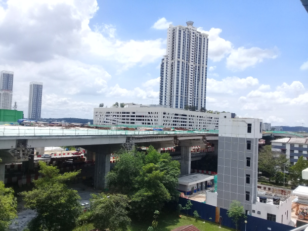 Luftaufnahme einer Stadtlandschaft mit Gebäuden, Bäumen, Fahrzeugen, einer Brücke und einer Baustelle für einen neuen Wohnkomplex unter einem bewölkten Himmel.
