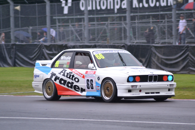 Ein weißes Rennauto mit Text und Zahlen darauf fährt auf einer Strecke, umgeben von Gras, mit Zuschauern hinter einem Maschendrahtzaun, einige halten Schirme, und ein Banner im Hintergrund.