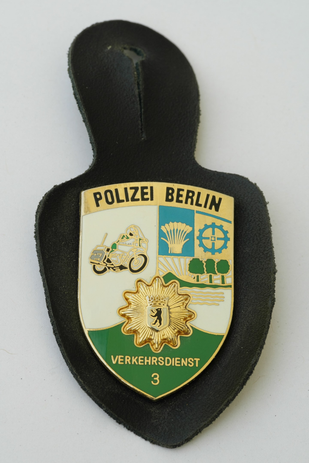 Eine Polizeimarke mit "Polizei Berlin" in fetter schwarzer Schrift auf einem blauen Schild mit einem weißen Stern und Rahmen.