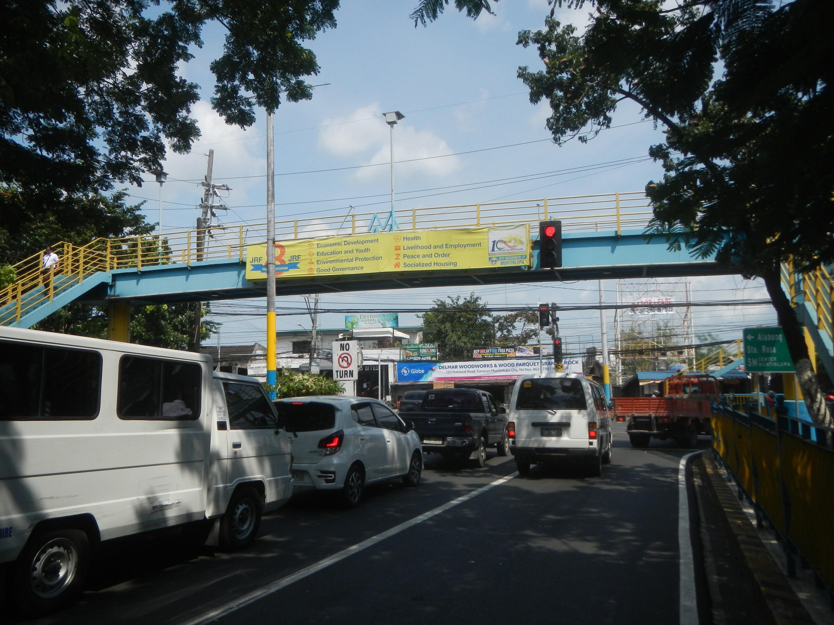 Vielbefahrene Straße mit Fahrzeugen, eine Brücke mit Geländern und Treppen, Laternen, Ampeln, Informationsschilder, Bäume, Gebäude und ein bewölkter Himmel.