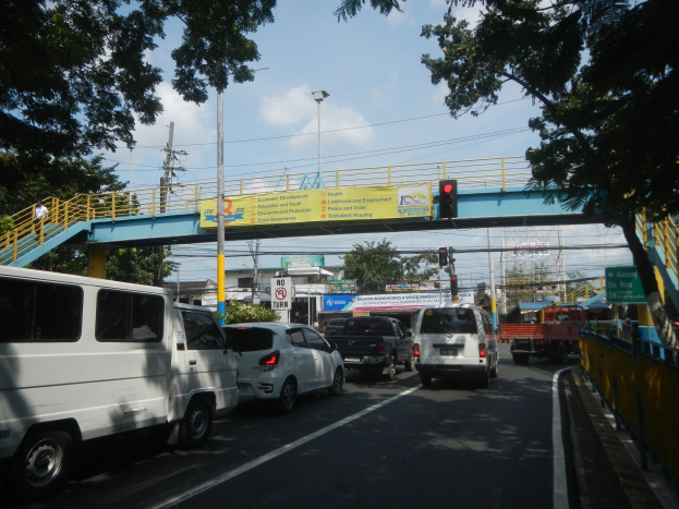 Vielbefahrene Straße mit Fahrzeugen, eine Brücke mit Geländern und Treppen, Laternen, Ampeln, Informationsschilder, Bäume, Gebäude und ein bewölkter Himmel.