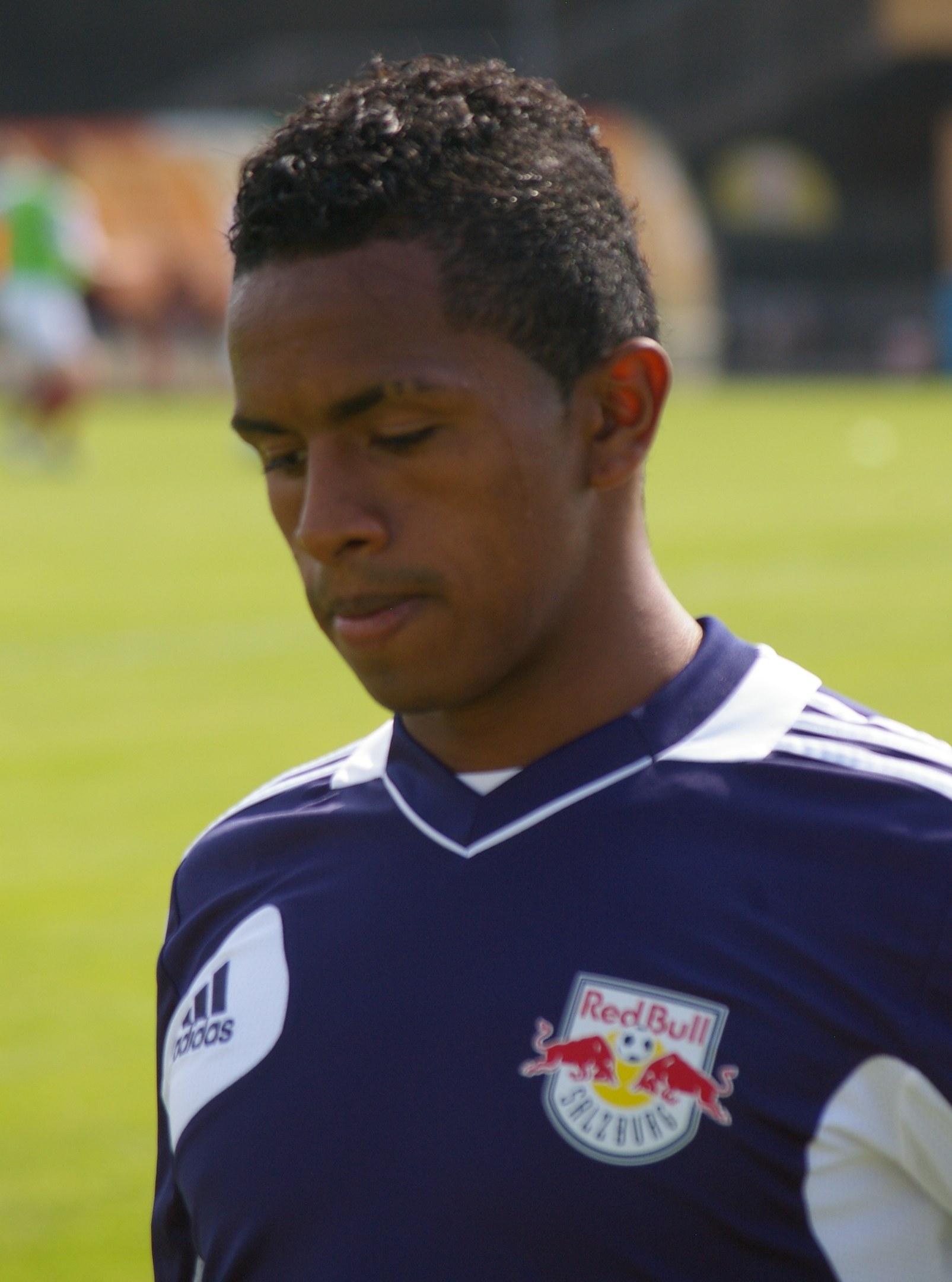 Junger Mann in blauem T-Shirt auf einem Fußballfeld mit unscharfem Hintergrund, der wie ein Flügelstürmer aussieht, mit kurzen dunklen Haaren und entschlossener Miene.