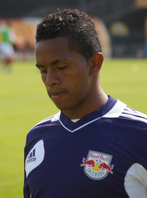 Junger Mann in blauem T-Shirt auf einem Fußballfeld mit unscharfem Hintergrund, der wie ein Flügelstürmer aussieht, mit kurzen dunklen Haaren und entschlossener Miene.
