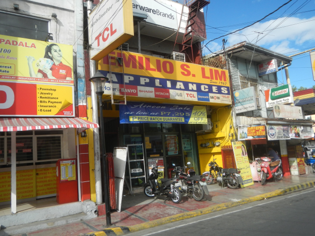 Eine belebte Straße mit parkenden Motorrädern, Strommasten, Bäumen und einem Geschäft mit der Aufschrift "Wilio S Lim Elektrogeräte" unter einem bewölkten Himmel.