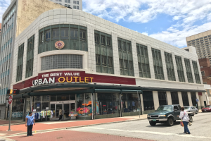 Eine belebte Stadtstraße mit Fahrzeugen, Fußgängern, hohen Glasgebäuden und einem Schild mit der Aufschrift "The Best Value Urban Outlet" unter einem bewölkten Himmel.