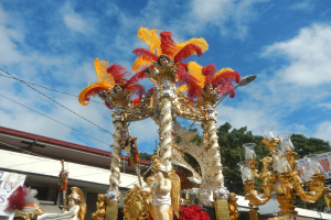 Ein Karnevalswagen mit goldenen und roten Federn, der Menschen trägt, wird in einem Umzug mit einem Gebäude, Bäumen, Drähten und einem Banner in der Nähe unter einem sichtbaren Himmel gezeigt.