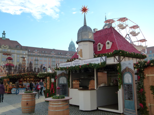 Ein geschäftiger Weihnachtsmarkt in Nürnberg, Deutschland, mit Menschen um dekorierte Buden, festliche Lichter, Schmuck, einem Riesenrad im Hintergrund, Gebäuden mit Fenstern und einem bewölkten Himmel.