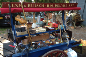 Ein blauer Karren voller Flaschen und Kartons auf einem Schotterweg auf einem Flohmarkt, mit Fahrzeugen, Bäumen und einer Wand im Hintergrund.