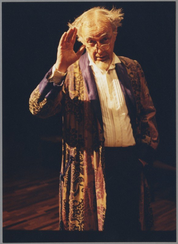 Ein Mann in einem langen Gewand und Brille steht auf einer Bühne mit gefalteten Händen und schaut auf ein dunkles Publikum.