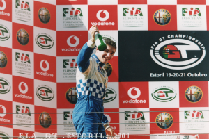 Ein Mann steht auf einem Rennpodest, hält eine Champagnerflasche in der Hand und lächelt, mit einem Hintergrund aus Logos und Text und Seilen am unteren Bildrand.