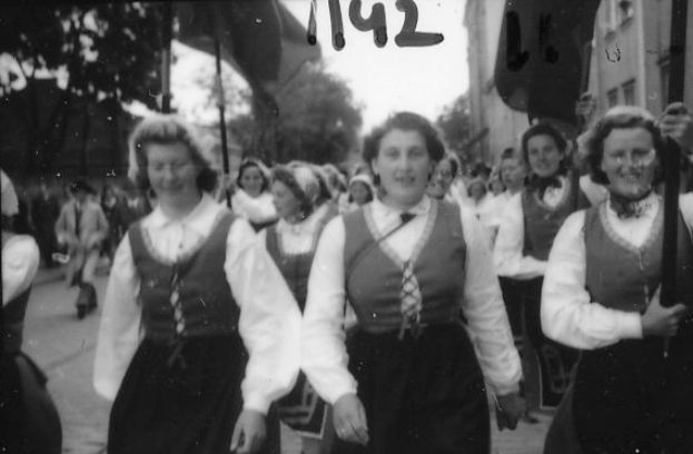 Eine Gruppe von Frauen in traditioneller bayerischer Tracht spaziert durch eine Straße, lacht und ist von Bäumen und Gebäuden umgeben, dargestellt in Schwarz-Weiß.