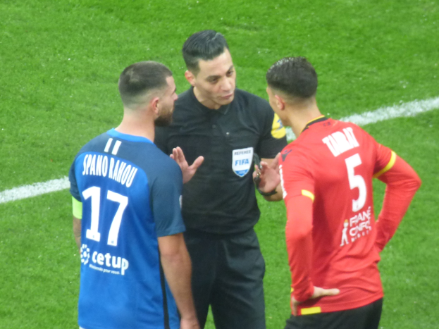 Schiedsrichter spricht mit zwei Männern auf einem Fuballfeld; einer in einem schwarzen Hemd und Hose, der andere in einem blauen und roten T-Shirt mit Text, beide tragen Brillen.